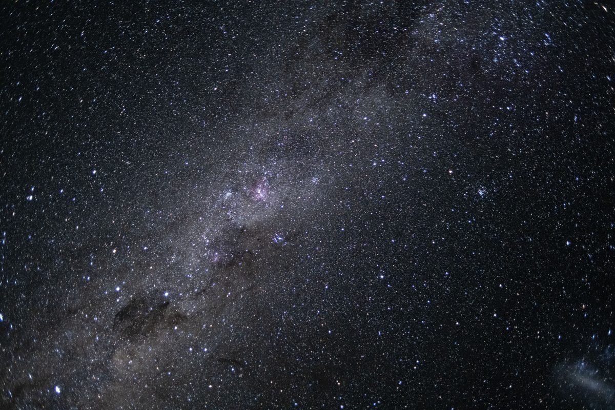 A dense band of stars and dark dust lanes of the Milky Way against black space, with a pink glowing nebula near the center of the frame